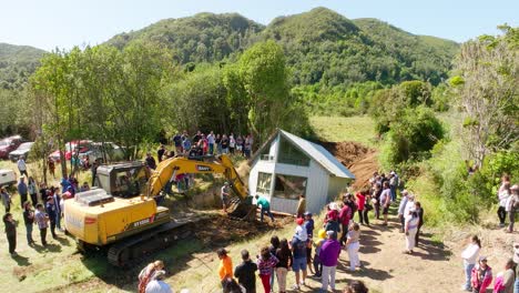 A-large-group-of-people-gathered-to-witness-the-relocation-of-a-house-aided-by-heavy-machinery-in-a-rural,-lush-green-landscape