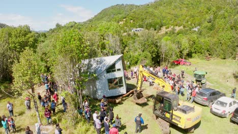 Vista-Aérea-De-Un-Gran-Grupo-De-Personas-Reunidas-Para-Presenciar-La-Reubicación-De-Una-Casa-Con-La-Ayuda-De-Maquinaria-Pesada-En-Un-Paisaje-Rural-Y-Verde-Y-Exuberante.