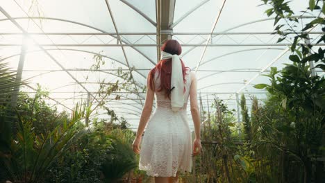 Filmische-Aufnahme-Einer-Jungen-Frau-Im-Weißen-Kleid-Und-Mit-Verbundenen-Augen-Aus-Einer-Niedrigen-Kameraperspektive,-Die-Von-Hinten-Durch-Ein-Gewächshaus-Mit-Pflanzen-Läuft