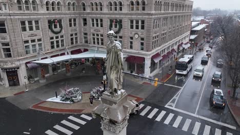 Toma-Aérea-En-órbita-De-Una-Estatua-En-El-Cruce-De-Una-Ciudad-Americana-Durante-Las-Nevadas-En-Invierno