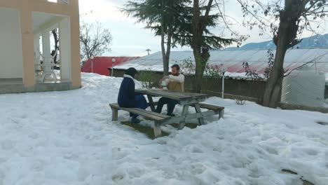 Two-individuals-are-engaged-in-conversation-or-an-activity-at-a-wooden-table-surrounded-by-snow,-with-trees-and-a-building-in-the-background