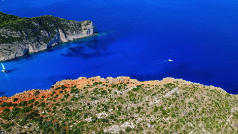 Aerial-drone-forward-moving-shot-over-sail-boats-entering-the-iconic-Navagio-Beach,-Greece-surrounded-high-cliffs-on-a-sunny-day