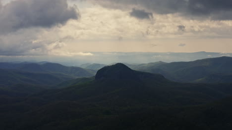 Sunset-aerial-of-large-rock-formation-towering-over-forest-in-mountains