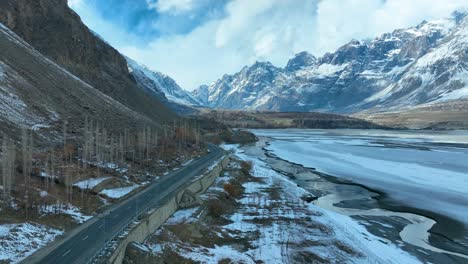 Un-Paisaje-Impresionante-Que-Muestra-Un-Camino-Sinuoso-A-Lo-Largo-De-Un-Río-Parcialmente-Congelado,-Flanqueado-Por-Montañas-Cubiertas-De-Nieve-Y-árboles-áridos-En-Skardu