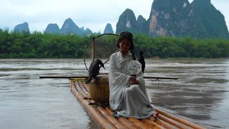 Hanfu-girl-smiles-while-seated-on-a-bamboo-raft,-holding-a-round-fan-with-cormorant-birds-beside-her