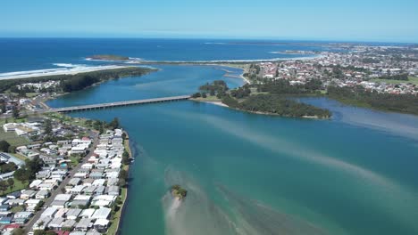 Panoramic-aerial-establishing-shot-of-Windang-with-coastal-homes-leading-to-bridge-and-strong-crashing-ocean-waves