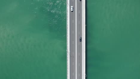 Drone-bird's-eye-view-tracking-follows-car-crossing-bridge-over-green-ocean-sea-water
