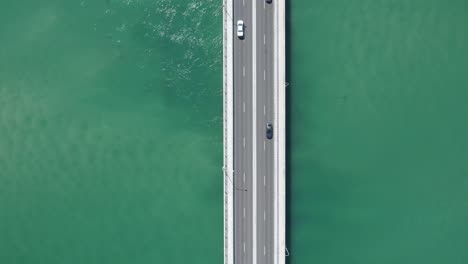 Die-Verfolgung-Durch-Die-Drohne-Aus-Der-Vogelperspektive-Folgt-Dem-Auto,-Das-Eine-Brücke-über-Das-Grüne-Meerwasser-überquert