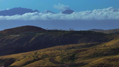 Stunning-dirt-road-winds-across-scenic-Maui-valleys-with-clouds-gathering-on-edge,-aerial-overview