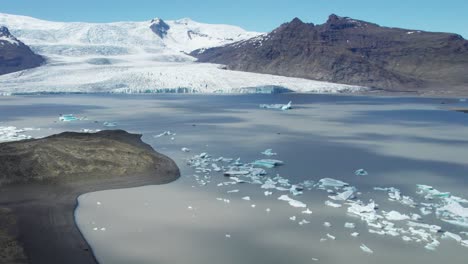 Luftaufnahme-Eines-Gletschersees-Mit-Schwimmenden-Eisbergen-Und-Dem-Vatnajokull-Gletscher-Im-Hintergrund-Im-Sommer,-Die-Die-Dynamische-Und-Atemberaubende-Naturlandschaft-Einfängt