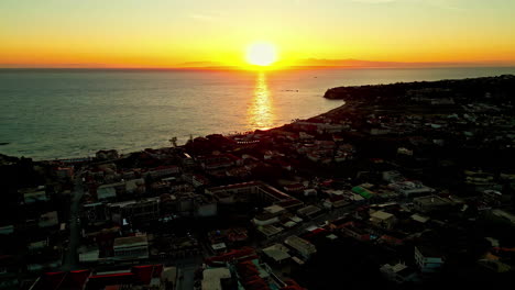 Drone-Aéreo-Hacia-Atrás-Disparado-Sobre-La-Ciudad-Costera-Con-Una-Increíble-Puesta-De-Sol-Dorada-En-El-Fondo-Durante-La-Noche