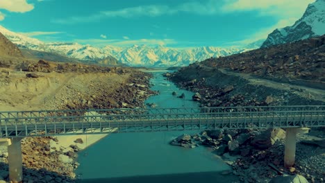 Aerial-view-capturing-a-bridge-spanning-a-blue-river-amidst-rugged,-rocky-terrain
