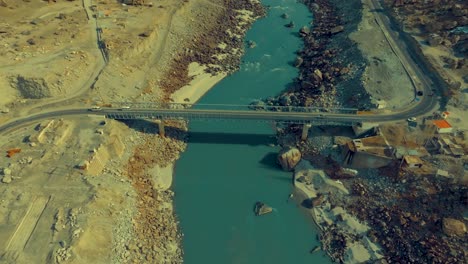 Aerial-view-capturing-a-bridge-spanning-a-blue-river-amidst-rugged,-rocky-terrain