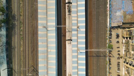 White-and-blue-blocked-pattern-on-top-of-Mumbai-station-as-train-waits-on-tracks,-aerial-top-down,-India