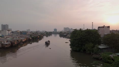 Vietnamesisches-Flussboot-Auf-Dem-Kanal-Mit-Städtischen-Hütten-In-Einem-Armen-Viertel-Von-Ho-Chi-Minh-Stadt,-Mit-Häusern-Und-Skyline-Im-Wunderschönen-Goldenen-Abendlicht