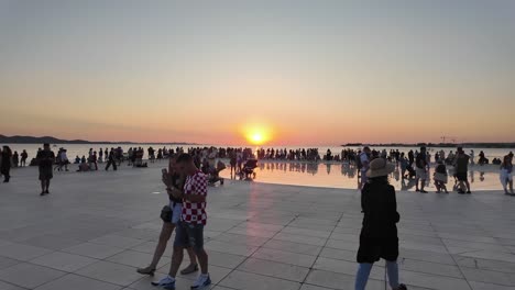 A-couple-wearing-Croatian-checkered-t-shirt-at-the-Greeting-the-sun-installation-in-Zadar,-passing-large-crowd-watching-sunset