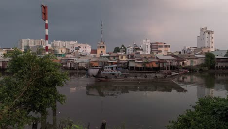 Vietnamesisches-Flussboot-Auf-Dem-Kanal-Mit-Städtischen-Hütten-In-Einem-Armen-Viertel-Von-Ho-Chi-Minh-Stadt,-Mit-Häusern-Und-Skyline-Im-Wunderschönen-Goldenen-Abendlicht