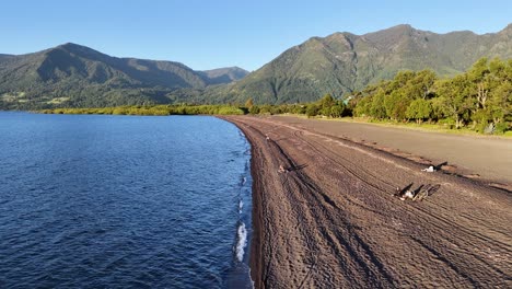 Low-flight-over-the-black-sand-on-the-shores-of-lake-Villarrica,-mountains-in-the-background