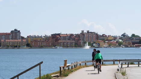 Pareja-De-Ancianos-Montando-En-Bicicleta-A-Lo-Largo-De-La-Pintoresca-Costa-De-Karlskrona,-Suecia---Toma-Amplia