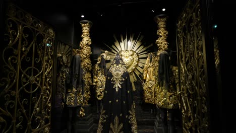 Dolce-Y-Gabbana,-Lujosas-Y-Ornamentadas-Figuras-Misteriosas-Con-Capas-Se-Alzan-Ante-Un-Corazón-Ornamentado-Decorado-En-Oro,-Creando-Una-Atmósfera-Solemne-Y-Teatral.