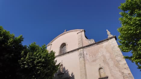 Vista-Exterior-Desde-Arriba,-Desde-La-Cruz-Hasta-La-Puerta,-La-Iglesia-De-San-Juan-Bautista-En-Fayence,-Francia-Con-Peatones-Y-Mirada-Desde-La-Pequeña-Plaza.