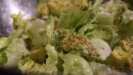 Summer-green-salad-with-lettuce,-avocado,-sesame-seeds-adding-olive-oil,-in-a-metal-bowl