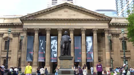 Die-Große-Fassade-Der-State-Library-Victoria-Mit-Der-Statue-Von-Sir-Redmond-Barry-Auf-Dem-Vorplatz,-Auf-Der-Sich-Einheimische-Und-Besucher-Auf-Der-Rasenfläche-In-Der-Innenstadt-Von-Melbourne-Aufhalten