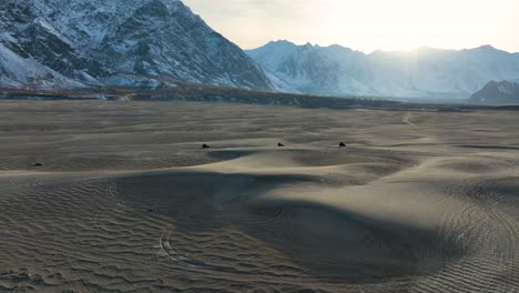 Vista-Aérea-Del-Paisaje-Desértico-Frío-De-Sarfaranga-Al-Atardecer-Con-Dunas-De-Arena-En-Primer-Plano-Y-Montañas-Con-Nieve-En-El-Fondo-Y-Vehículos-Cruzando