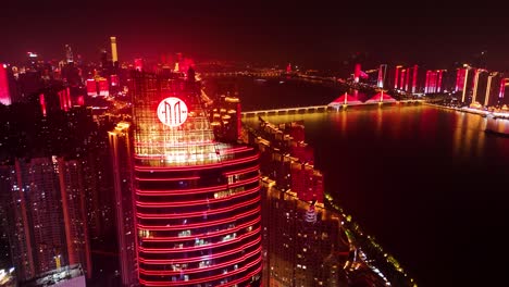 Vista-Nocturna-En-órbita-De-Un-Edificio-Comercial-Con-El-Lago-Y-El-Puente-Sanchaji-Al-Fondo-En-La-Ciudad-De-Changsha,-Hunan,-China