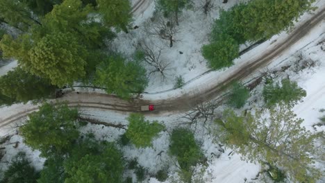 An-overhead-shot-captures-a-red-car-traversing-a-winding-road-surrounded-by-snow-covered-trees-and-terrain