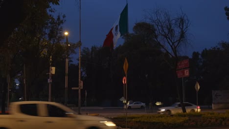 Busy-car-traffic-street-in-Mexico-City-at-night,-big-waving-Mexican-flag