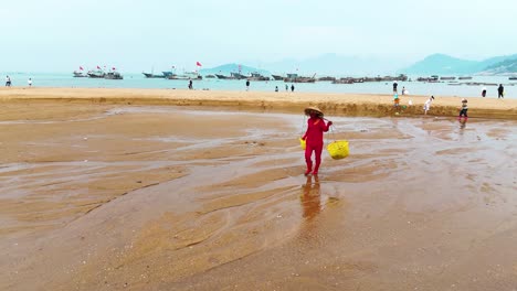 Una-Pescadora-Con-Cestas-Caminando-Por-La-Orilla-Con-Barcos-De-Pescadores-Y-Barcos-Al-Fondo