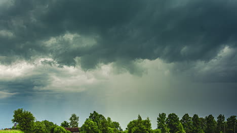 Dark-storm-clouds-roll-over-a-green-landscape-with-trees-swaying-in-the-wind,-timelapse