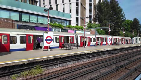 Un-Tren-Subterráneo-De-Londres-Se-Detuvo-En-Una-Plataforma-Con-Pasajeros-Y-El-Icónico-Logo-Circular-En-Una-Mañana-Soleada