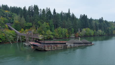 Drone-footage-of-a-barge-being-loaded-with-gravel-on-Puget-Sound,-surrounded-by-a-pine-forest-in-autumn