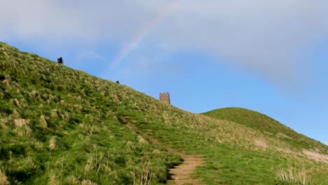 People-walking-up-to-The-Tor-in-Glastonbury-with-colourful-rainbow-over-the-ancient-tower-on-the-Isle-of-Avalon-in-Somerset,-England-UK