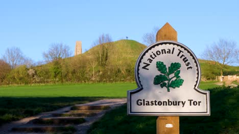 Scenic-landscape-view-of-The-Tor-with-signpost-for-National-Trust-in-rural-countryside-of-the-Somerset-Levels-in-Somerset,-England-UK