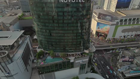 Top-down-aerial-view-of-Novotel-Bangkok-Platinum-hotel,-showcasing-an-open-balcony-and-terrace-café,-with-a-bustling-road-below-teeming-with-car-traffic,-highlighting-the-dynamic-and-lively-cityscape