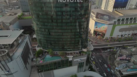 Vista-Aérea-De-Arriba-Hacia-Abajo-Del-Hotel-Novotel-Bangkok-Platinum,-Que-Muestra-Un-Balcón-Abierto-Y-Una-Cafetería-Con-Terraza,-Con-Una-Calle-Bulliciosa-Debajo-Repleta-De-Tráfico-De-Automóviles,-Resaltando-El-Dinámico-Y-Animado-Paisaje-Urbano