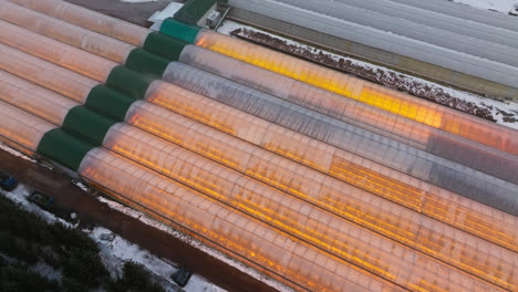 Drone-rotating-over-a-large-illuminated-greenhouse,-on-a-snowy,-winter-evening