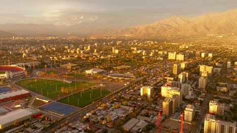 Hermosa-Vista-Aérea-Panorámica-De-Santiago-Al-Atardecer-Con-Un-Estadio,-Infraestructura-Urbana-Y-Montañas-Al-Fondo