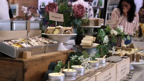 Homemade-soaps-for-sale-displayed-on-stall-during-market-fair-with-people-walking-and-looking