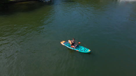 Woman-Sits-with-Dog-on-Paddle-Board-in-Lake