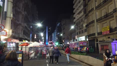 Argentine-people-walk-at-night-in-Corrientes-Avenue,-commercial-city-center-with-buses-and-obelisk-background-of-buenos-aires,-vibrant-latin-american-Capital