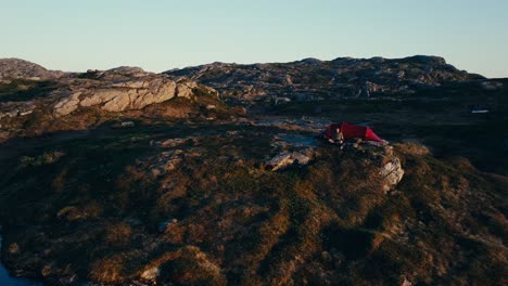 A-Man-Sits-Next-to-His-Red-Tent-Set-Up-on-the-Rugged-Terrain-in-Skurven,-Eidsvoll,-Akershus,-Norway---Orbit-Drone-Shot