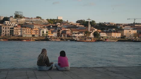 Two-people-sitting-by-the-Douro-River,-overlooking-the-Porto-skyline-at-sunset,-Portugal