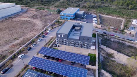 aerial-drone-shot-of-a-square-building-on-metal-stilts-with-solar-panels-and-cars-parked-below-and-on-the-side-of-the-road-to-the-left