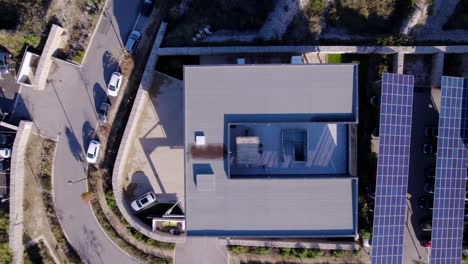 aerial-drone-shot-of-a-square-building-with-solar-panels-lined-up-on-the-parking-lot-and-cars-parked-underneath