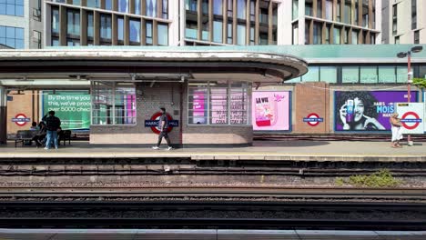 Los-Viajeros-Esperando-En-Una-Grada-En-El-Andén-De-Una-Colina-En-Una-Estación-De-Tren-Con-Anuncios-Y-Carteles