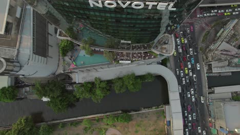 Top-down-aerial-of-Novotel-Bangkok-Platinum-hotel,-featuring-an-open-balcony-and-terrace-café,-with-a-busy-road-below-filled-with-car-traffic,-illustrating-the-concept-of-urban-and-vibrant-city-life
